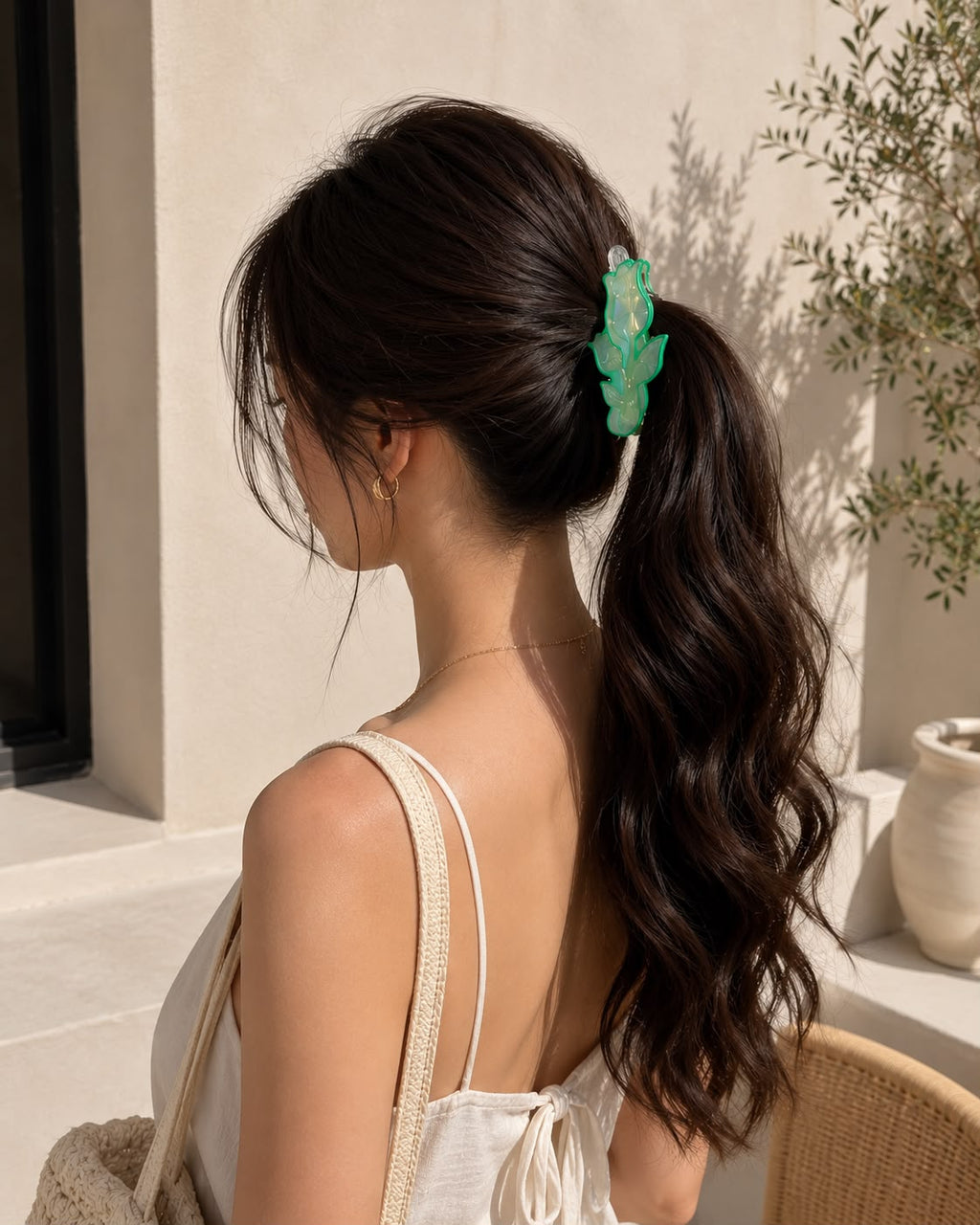 A woman shown from the back wearing a light green leaf-shaped hair claw clip that holds her dark brown wavy hair in a ponytail, set against a warm beige wall background.