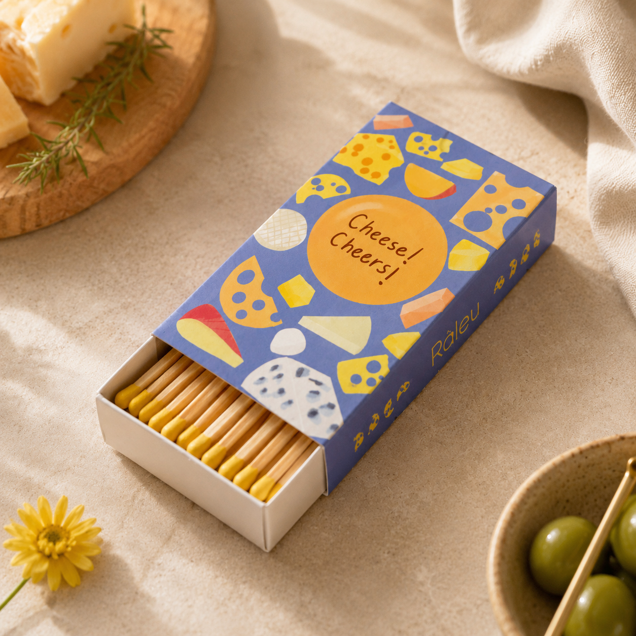 A box of Raleau cheese-themed crackers sits open on a beige fabric surface, surrounded by styled food props including cheese wedges, olives, and a small yellow flower. The box lid is decorated with colorful illustrations of cheese and the words 'Cheese! Cheers!'.